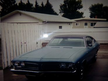 Classic blue vintage car parked in a driveway with white fences and houses in the background.