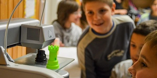 Niños observando una impresora 3D imprimiendo una torre de ajedrez verde.