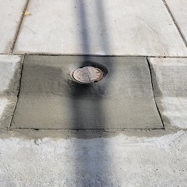 Freshly poured concrete patch around a manhole on a sidewalk.