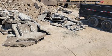 Piles of broken concrete slabs near a black utility trailer on a dirt surface.