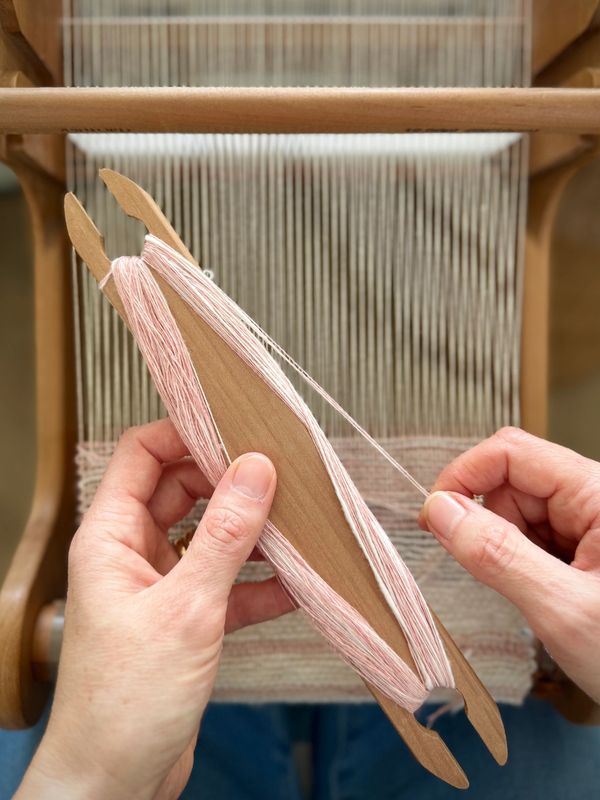 Hands winding soft pink thread on a wooden weaving shuttle near a loom.