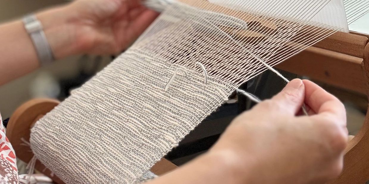 Hands weaving white thread on a wooden loom.