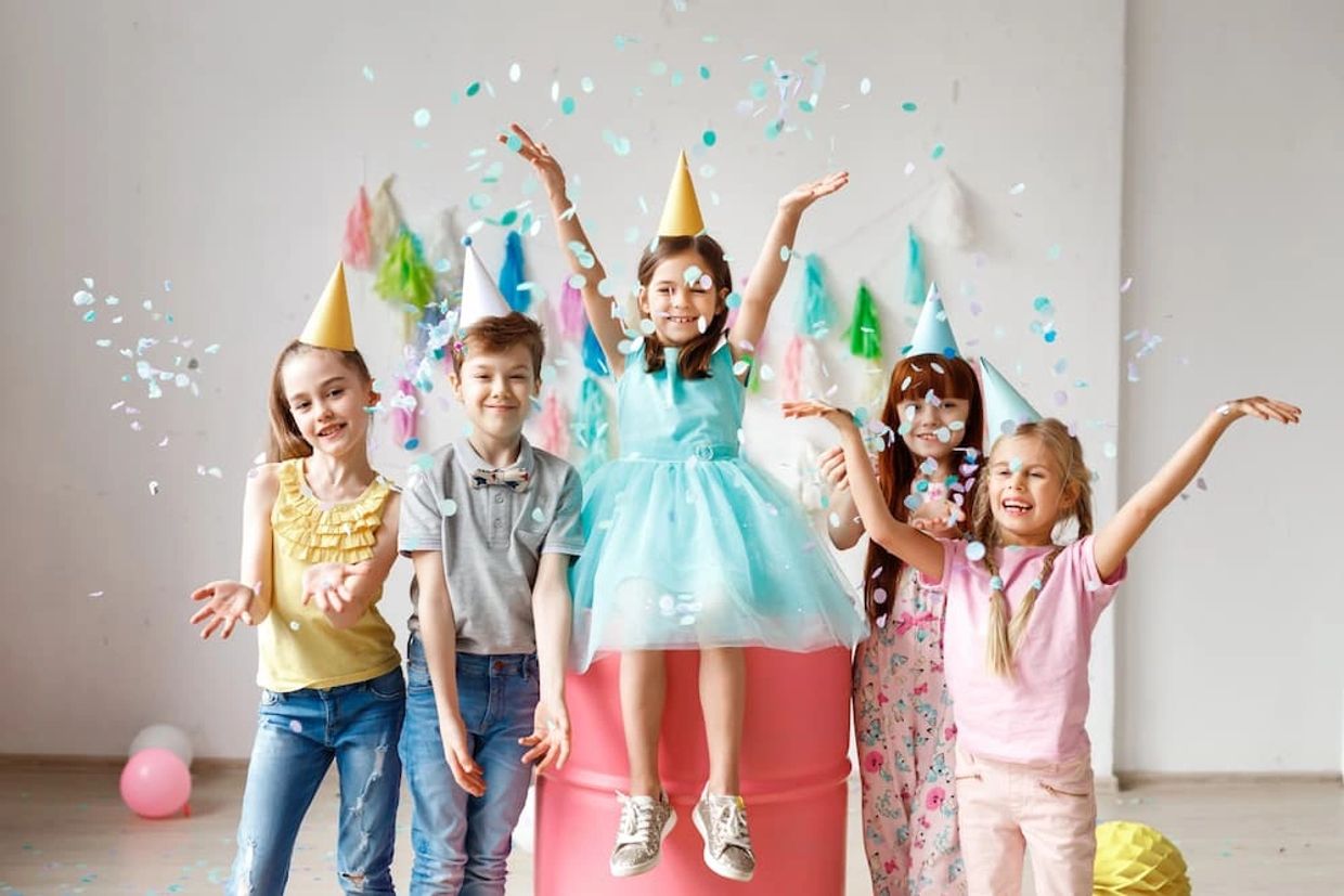 Five children wearing party hats celebrating with confetti indoors.