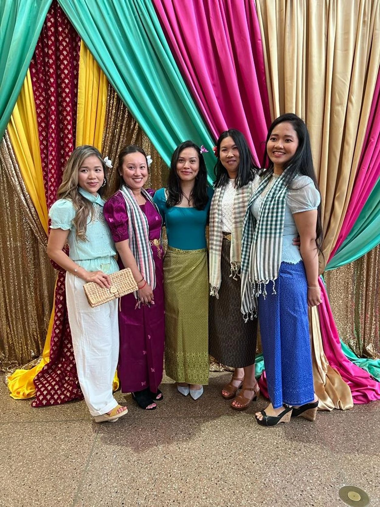 Five women in traditional attire posing in front of colorful draped fabric.