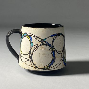 White ceramic mug with fluid circular design. Cool color accents. Black handle, base, interior.