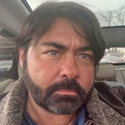 Man with dark hair and beard sitting inside a car.