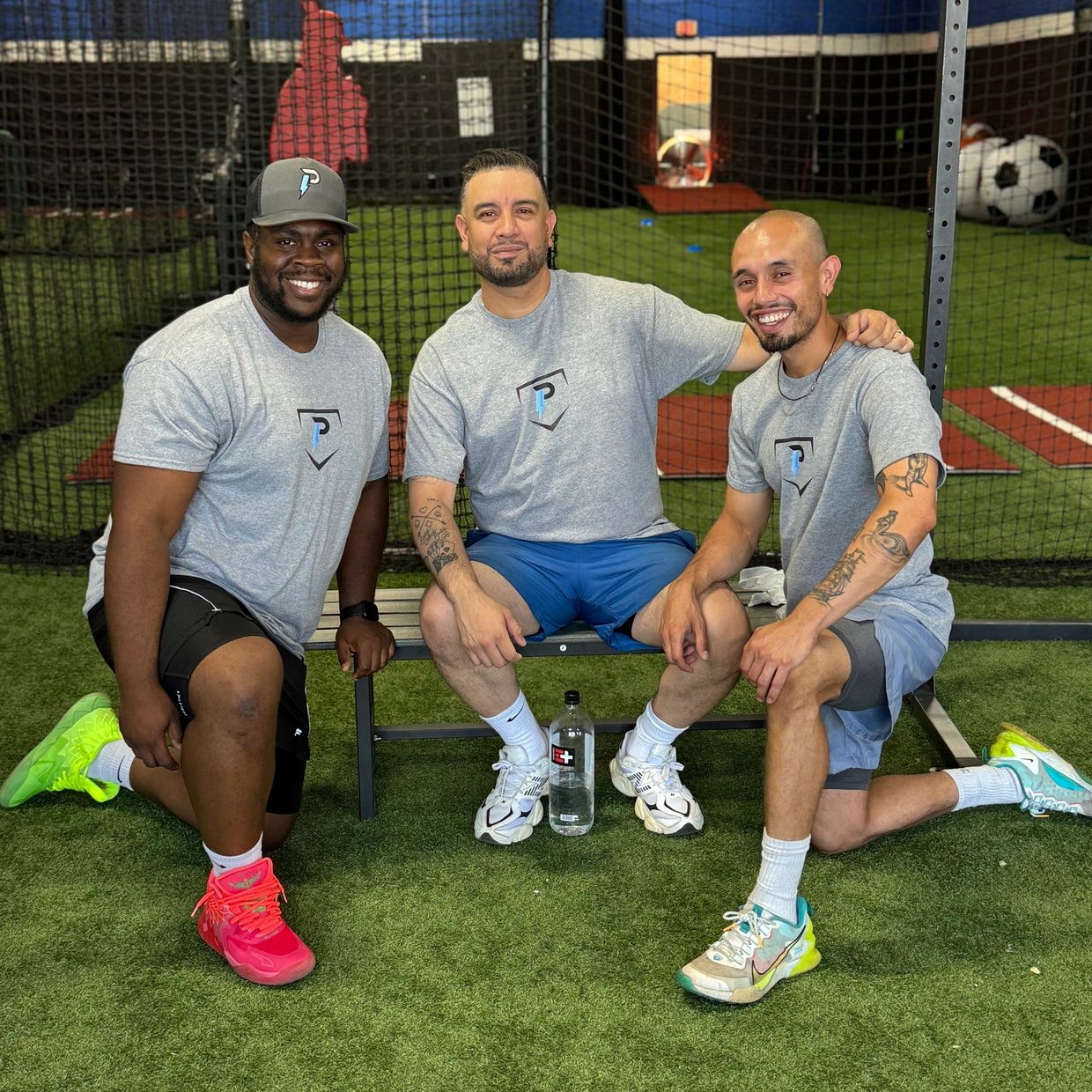 Three men in matching gray shirts posing indoors on artificial turf.