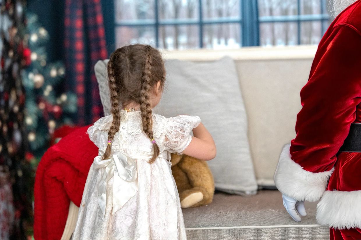 Little girl with braided hair wearing white dress near Santa Claus.