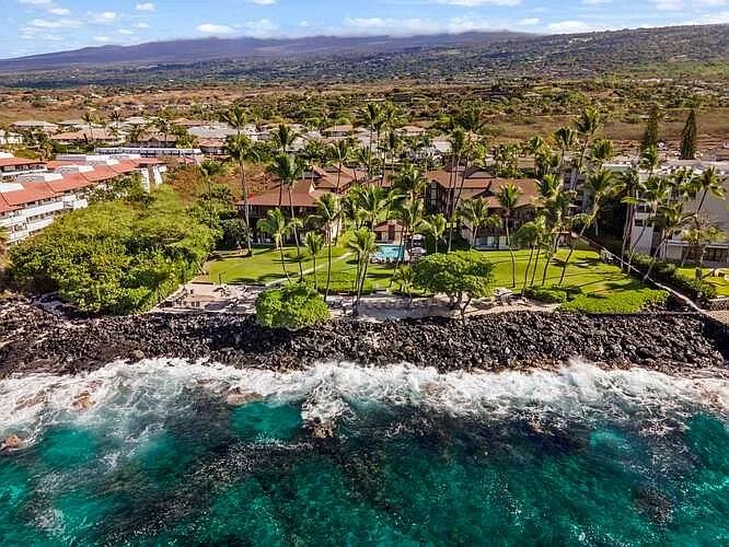 Oceanfront resort with palm trees and rocky shore under a blue sky.