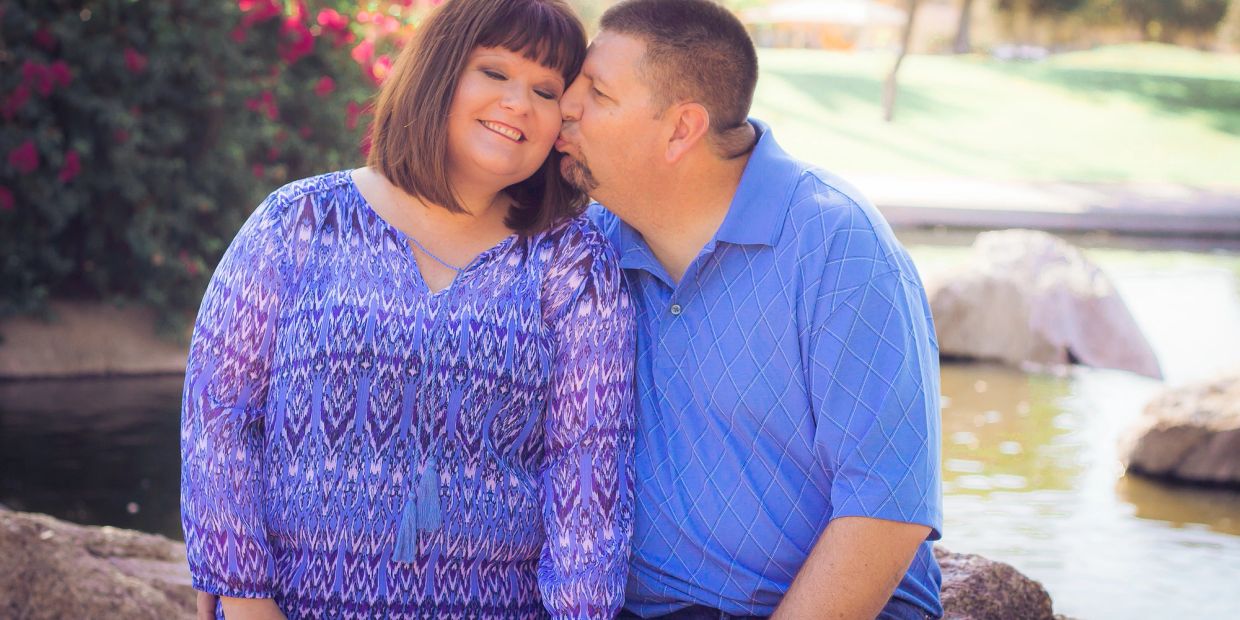 A man kisses a smiling woman on the cheek by a peaceful pond.