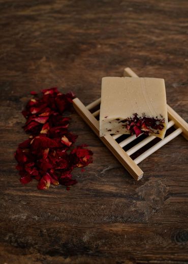 A bar of soap with rose petals on a wooden soap dish beside dried rose petals.