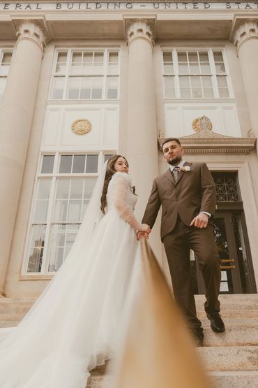 Newlyweds pose on grand steps of a historic building, holding hands along a railing.