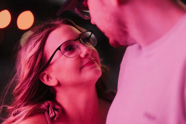 Woman with glasses gazes lovingly at a man under soft pink lighting.
