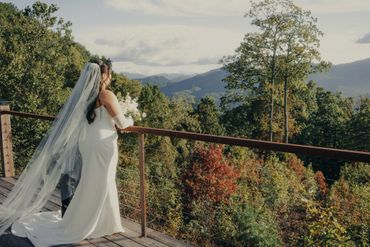 Bride in white gown overlooking scenic mountain landscape from balcony.