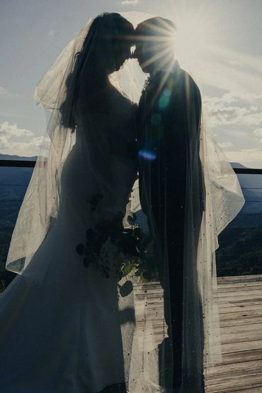 Silhouetted bride and groom tenderly touching foreheads with sun flare behind them.