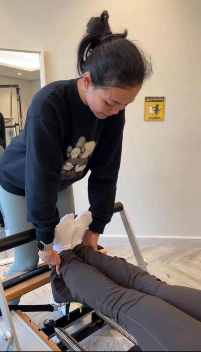 A woman assists another with leg exercises on a Pilates reformer machine.
