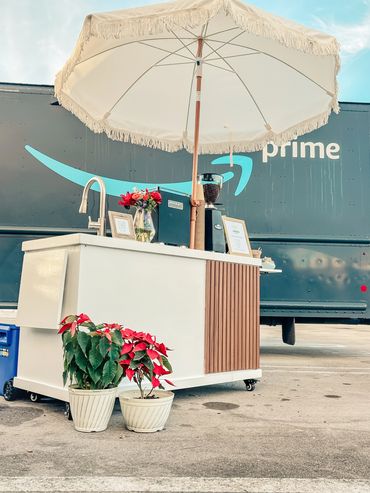 A stylish mobile coffee cart with umbrella and plants in front of an Amazon Prime truck.