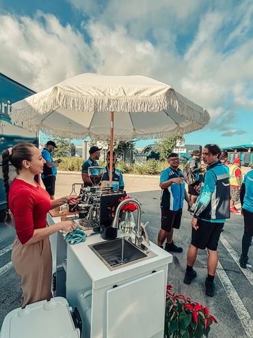 A woman prepares coffee at an outdoor stand with people socializing nearby.