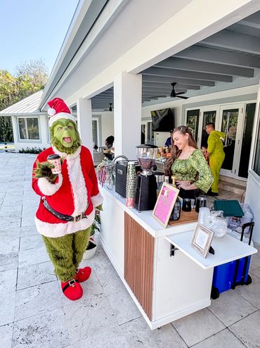 Person dressed as the Grinch in a Santa suit holding a coffee cup at an outdoor bar.