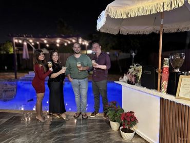 Four friends enjoy drinks by a poolside bar under night lights.
