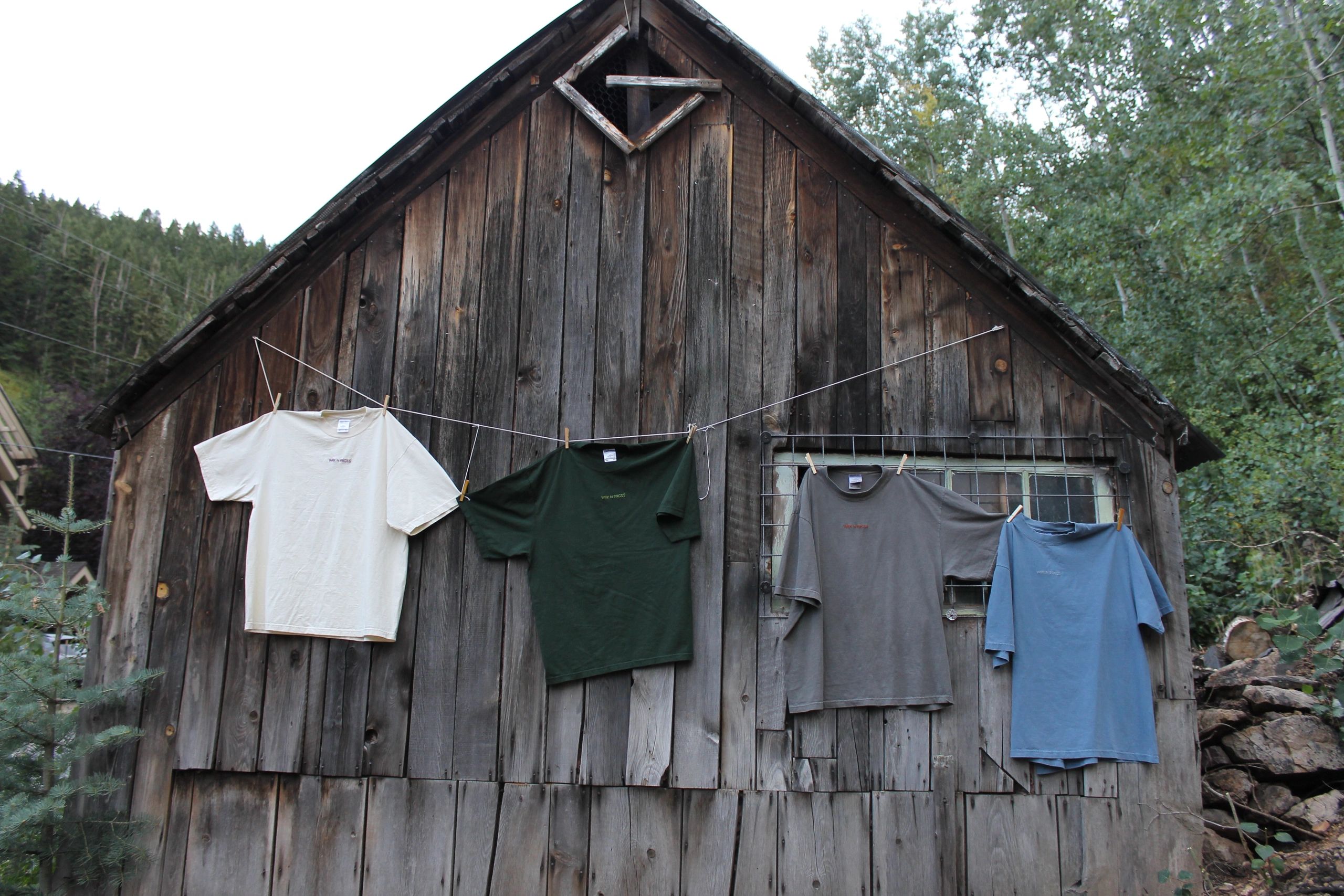Four t-shirts hanging on a clothesline outside a rustic wooden cabin.