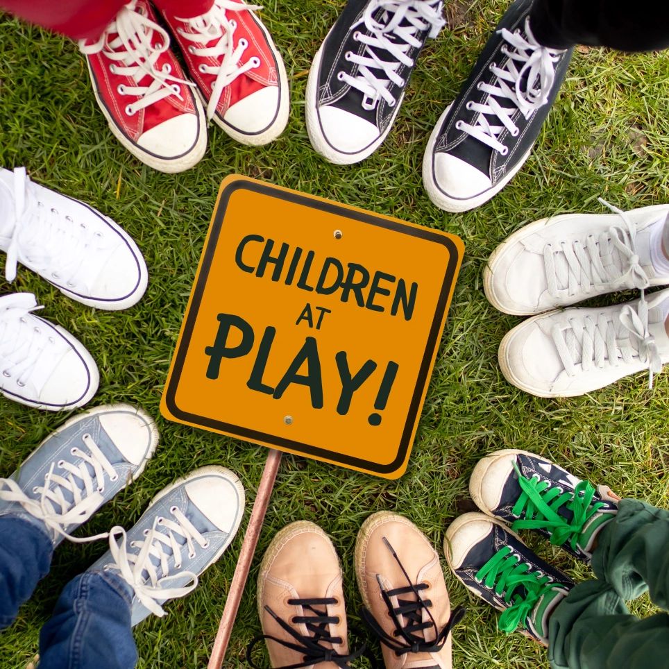 Children's colorful sneakers surround a 'Children at Play!' sign on grass.