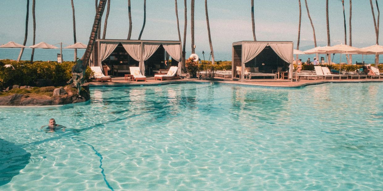 A person swimming in a large pool with cabanas and palm trees.