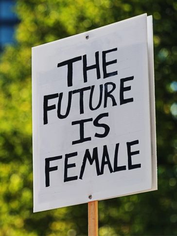 A sign reads 'THE FUTURE IS FEMALE' against a blurred green background.