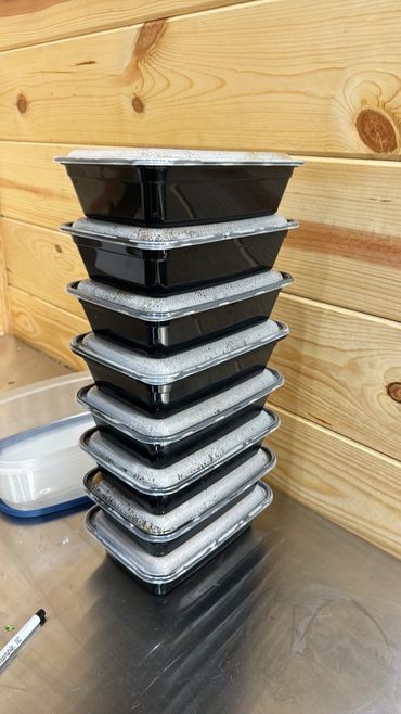 Stack of black food containers with sealed lids on a metal surface.