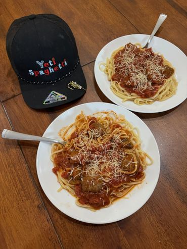 Two plates of spaghetti with tomato sauce and cheese next to a black cap on a wooden table.