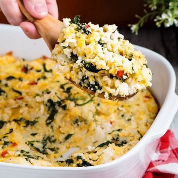 A hand scoops cheesy spinach rice casserole from a white baking dish.