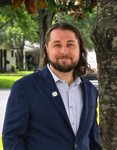 Man in blue blazer standing by a tree outdoors.