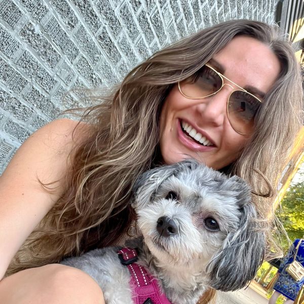 Smiling woman in sunglasses holding a small dog wearing a pink harness.