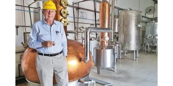 Elderly man in a hard hat standing in a distillery with copper and stainless steel equipment.