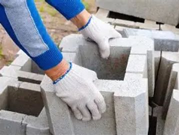 Hands wearing gloves handling concrete blocks outdoors.