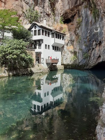 White house built against a rocky cliff, reflected in calm water.