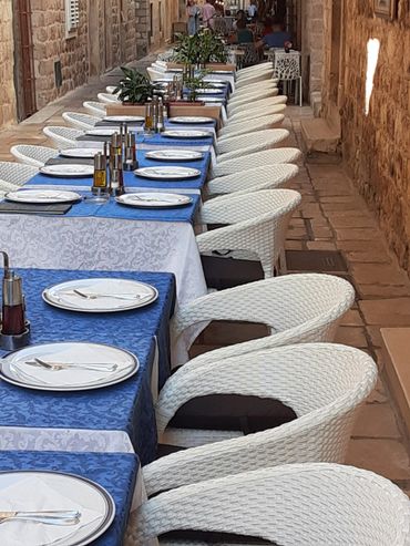 Outdoor dining setup with white wicker chairs and blue tablecloths along a stone wall.