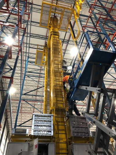 Workers operate a blue lift near a tall yellow industrial structure inside a warehouse.