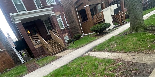 A sidewalk between brick houses with green lawns and trees.