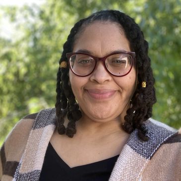 Smiling woman with glasses and braided hair outdoors.