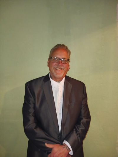 Smiling man in a black suit and glasses standing against a plain wall.