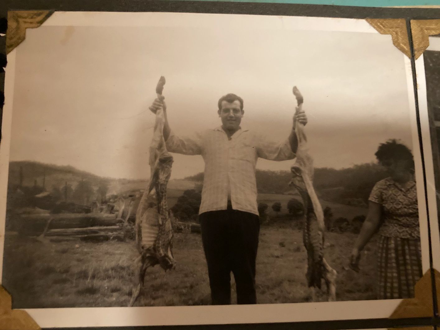 Vintage photo of a man holding two skinned animals outdoors, with a woman standing nearby.