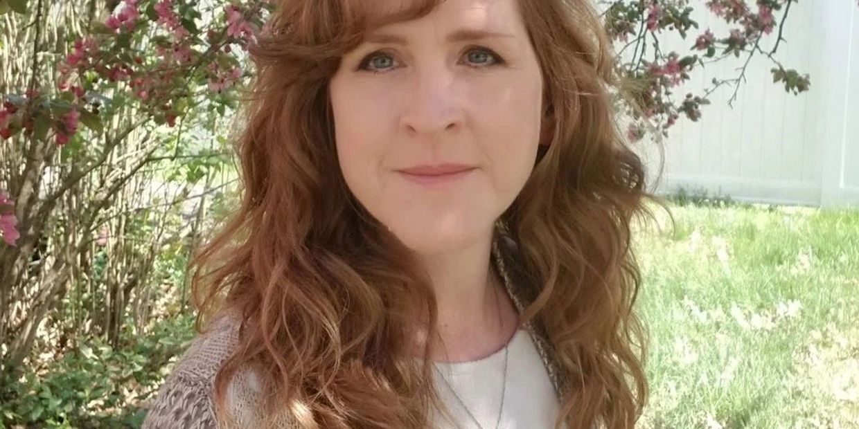 Woman with red curly hair stands outdoors near flowering bushes.
