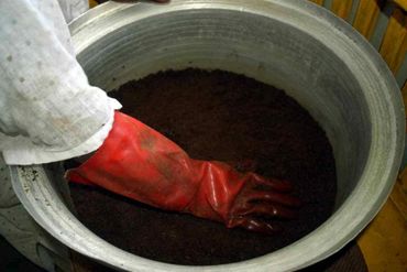 A hand in a red glove touching dark soil in a large metal container.
