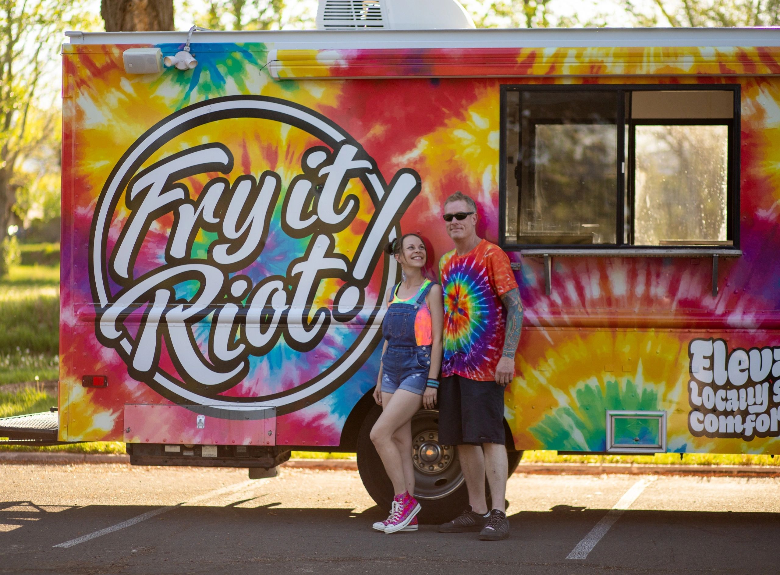 Two people stand smiling in front of a colorful tie-dye food truck named Fry it Riot. Book us today