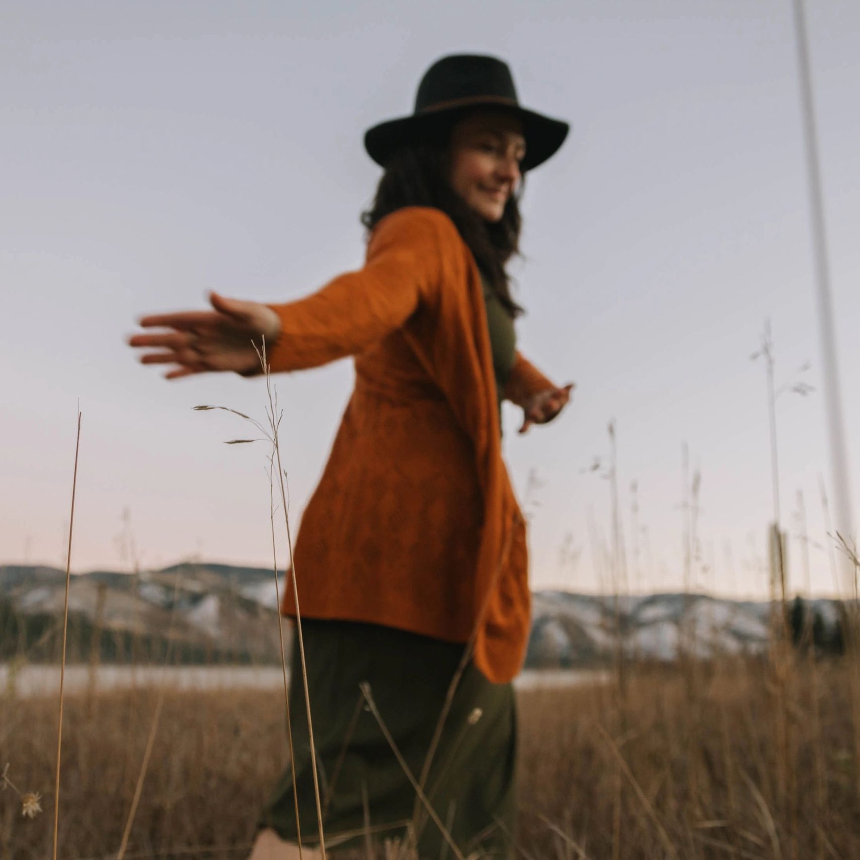 A woman in a hat and orange sweater walks through a field at dusk. Through heart activation,  and mindful movement she experiences Divine love and personal sovereignty.