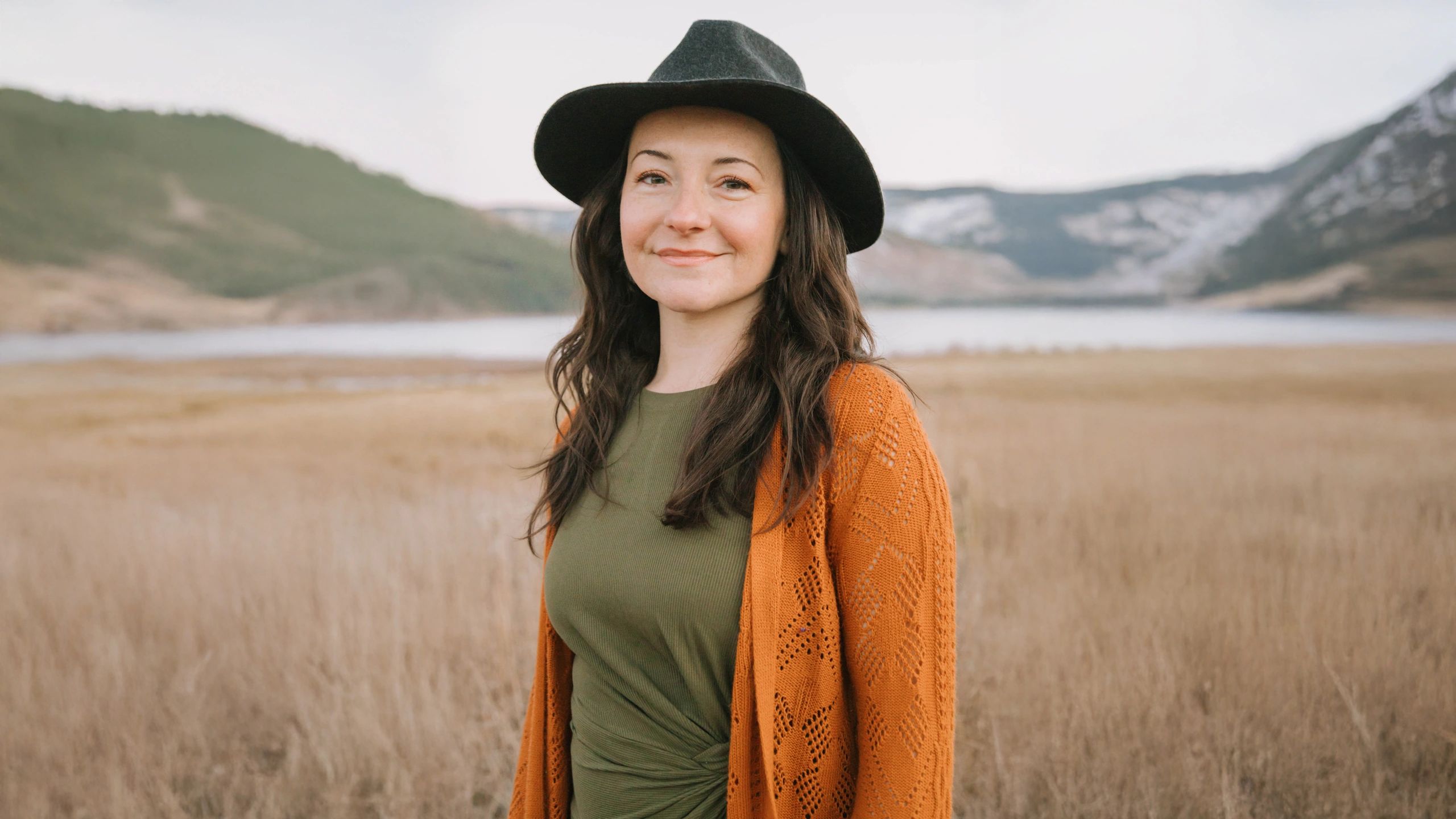 Woman smiling in a field wearing a black wide-brimmed hat and orange cardigan. She has a peaceful presence and a quiet knowing in her gaze. She understands how heart activation can transform your life.