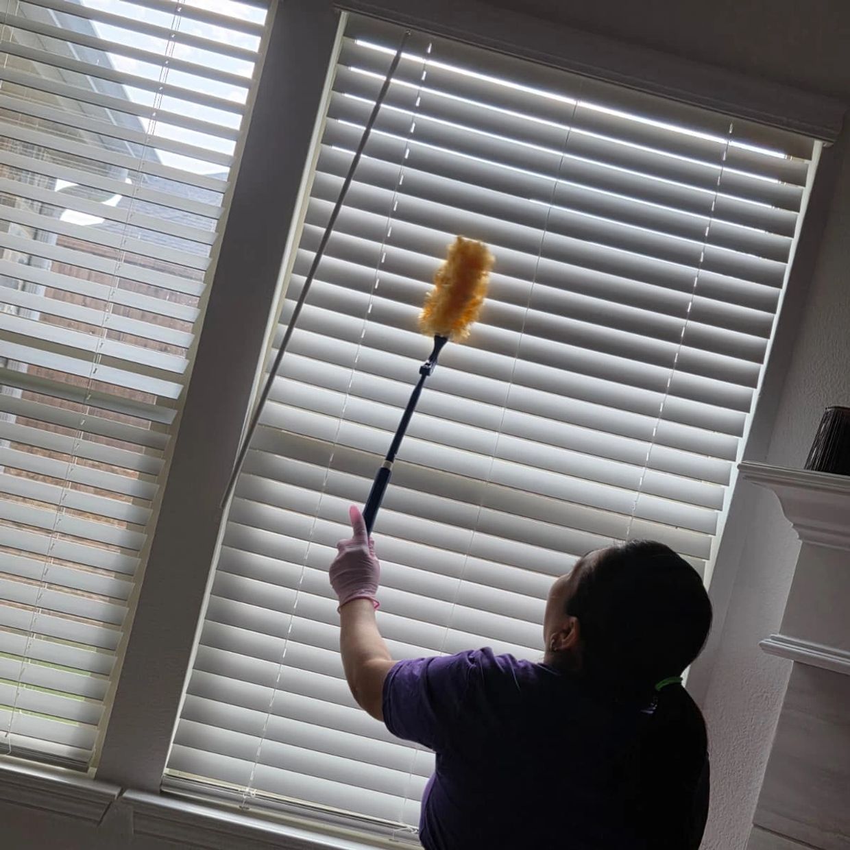Person dusting white window blinds with a yellow duster.