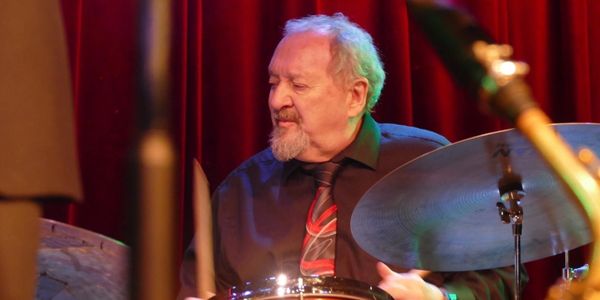 An elderly man passionately playing drums on stage with red curtains behind.