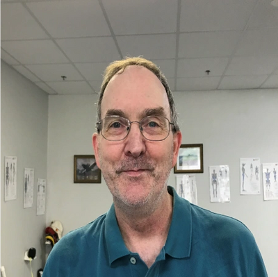 Middle-aged man with glasses in a teal polo shirt, smiling slightly in a clinical room.